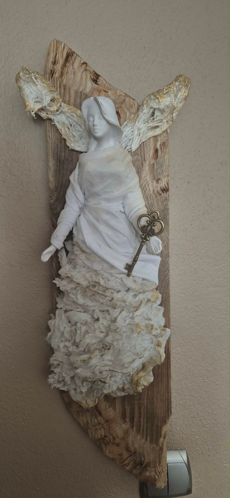White plaster bust of a woman with gold leaf wings mounted on weathered driftwood, holding an ornamental key