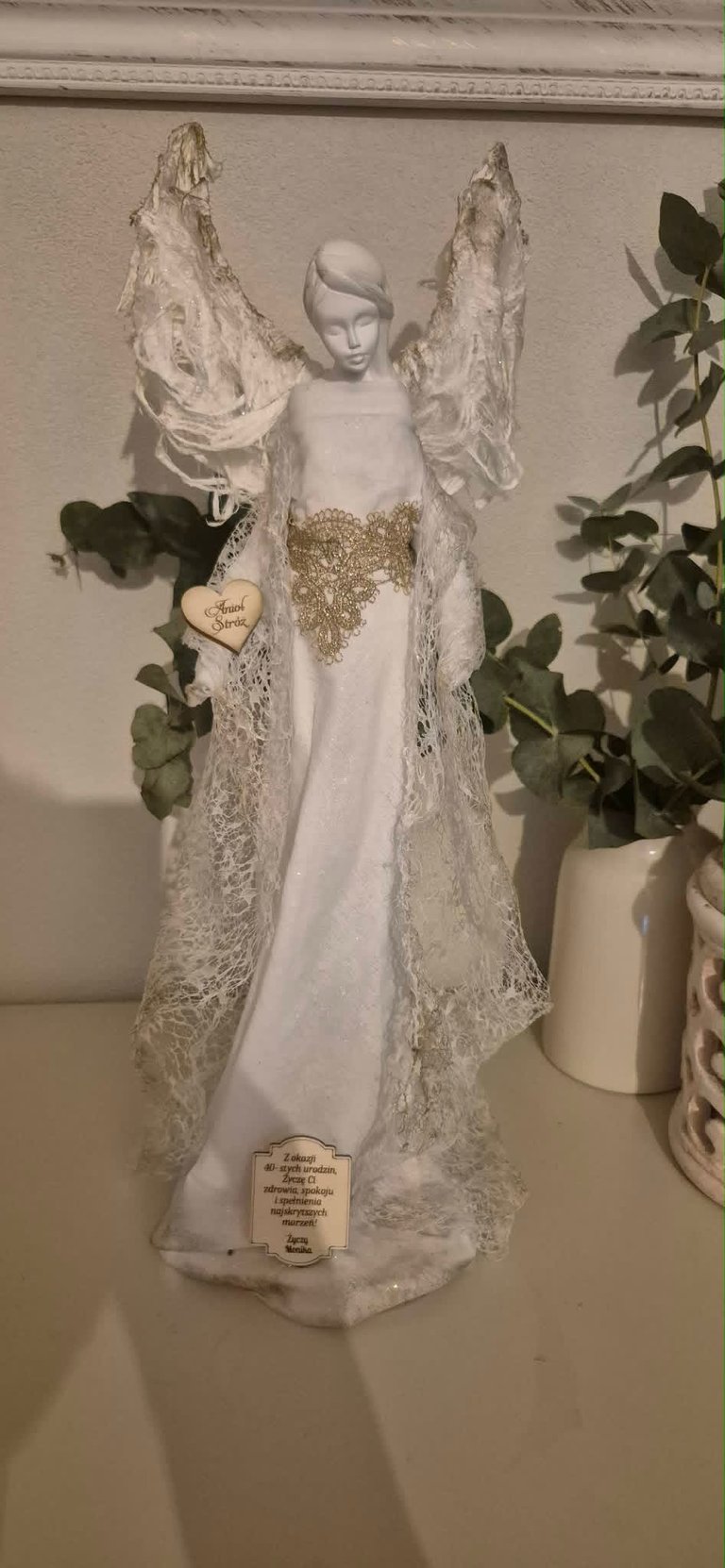 White decorative angel figurine with lace wings and gold accents, standing among potted plants on a shelf