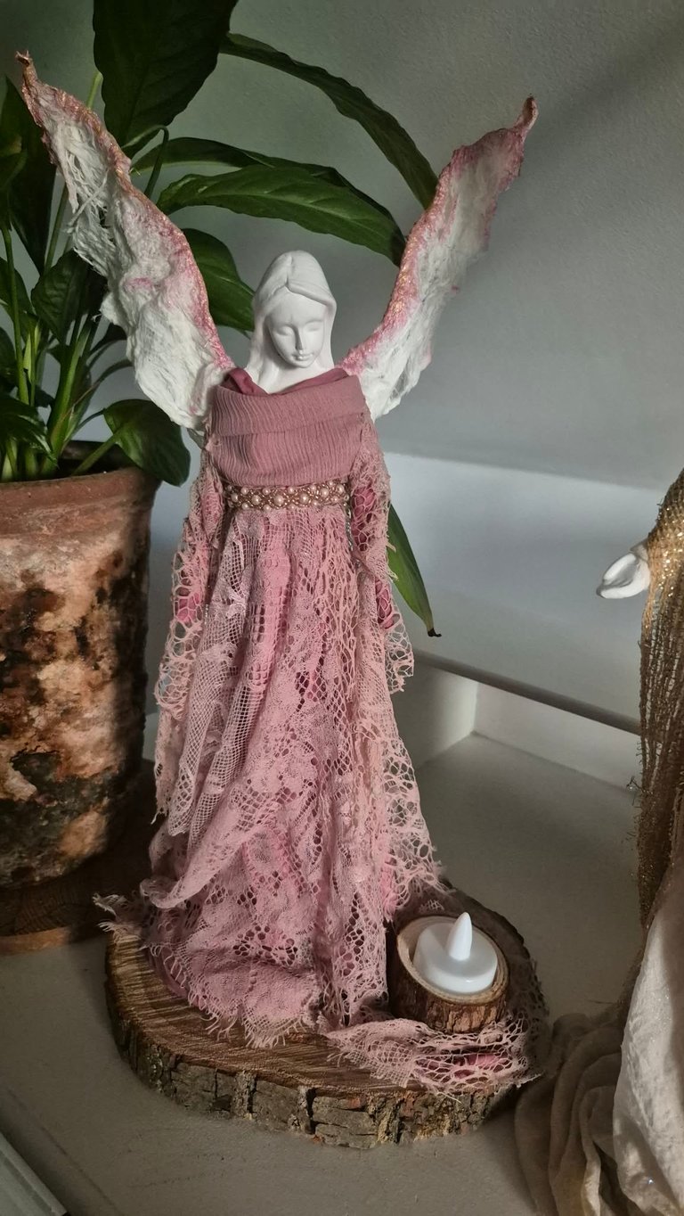 Decorative angel figurine with white head and pink lace dress and wings on wooden base, surrounded by potted plants
