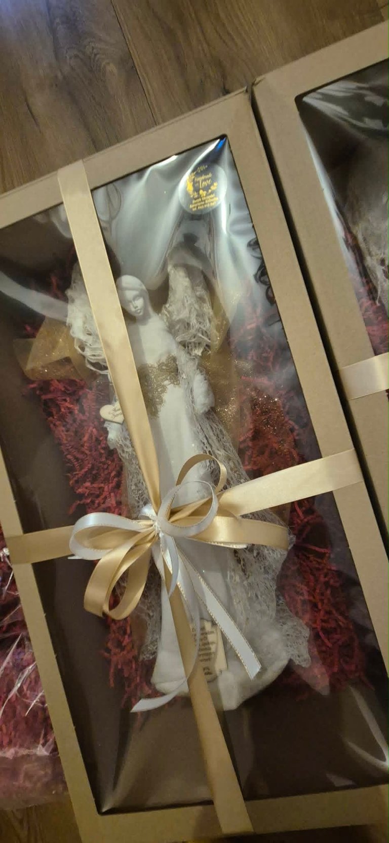 Packaged decorative angel figurine with white dress and gold accents, tied with cream ribbon bow, displayed in tan box on wooden floor