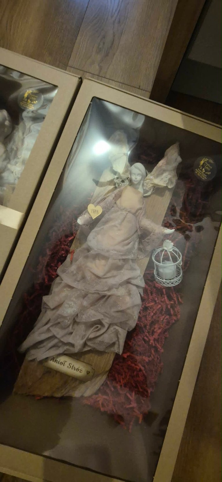 Religious statue in glass display case with angel figurine, candle, and red decorative elements on wooden floor
